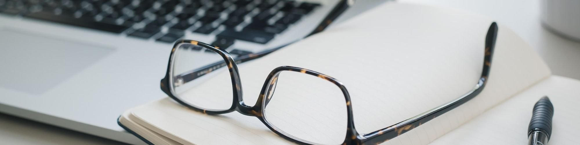 Glasses on a desk with a laptop computer and books.